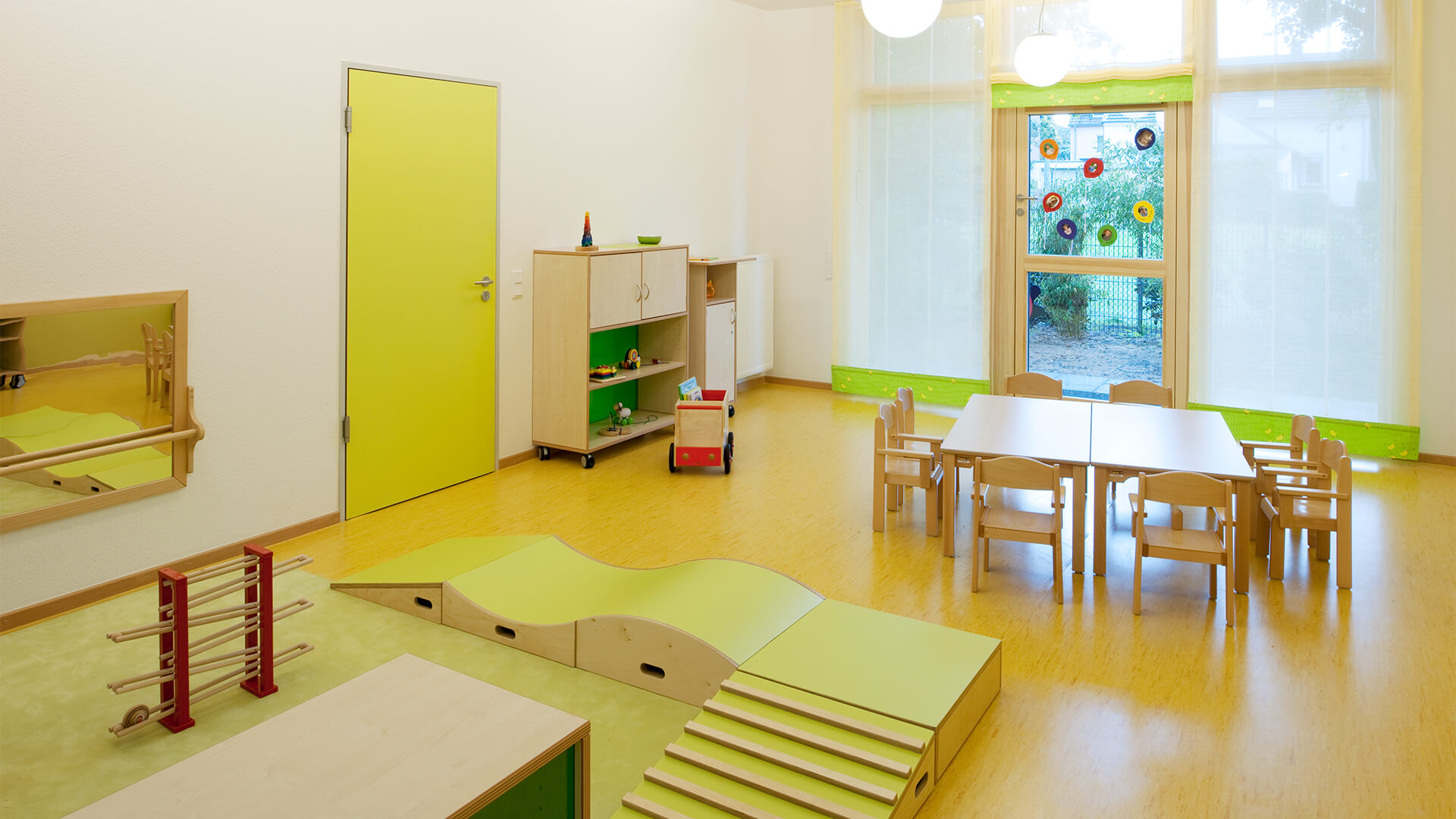 Gruppenraum der Kindertagesstätte in Neuss mit Tischen und Spielecke auf grünem Teppich